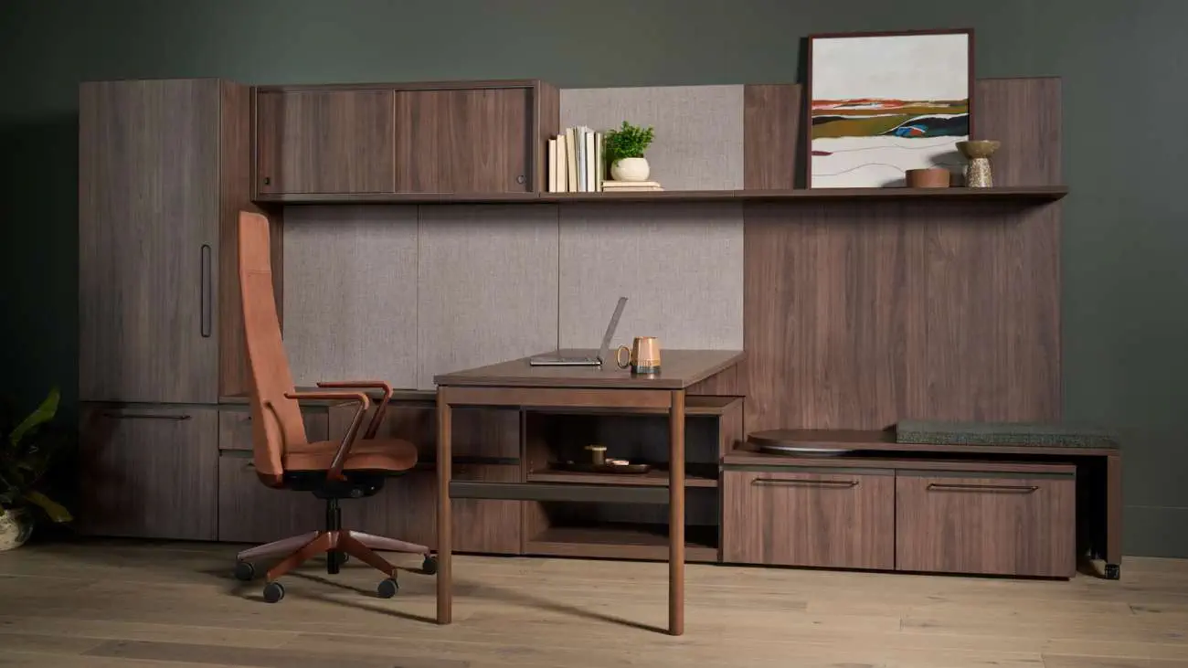 OFS Adler desk in walnut finish with matching wall-mounted storage cabinets and shelving, paired with a tan leather swivel chair in a modern office setting.